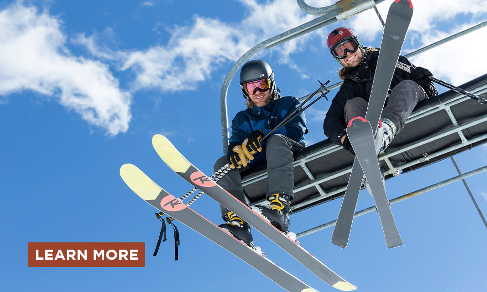 Two skiers in a gondola looking down at the camera with blue sky and a Learn More button.